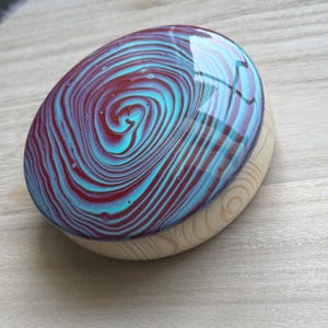May include: Oval wooden box with a swirling pattern in teal and burgundy. The lid has a glossy finish, and the base is made of light-colored wood. The design resembles a galaxy or abstract art.