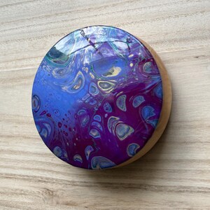 May include: A round wooden coaster with a purple, blue, and gold abstract design. The coaster is covered in a clear epoxy resin.
