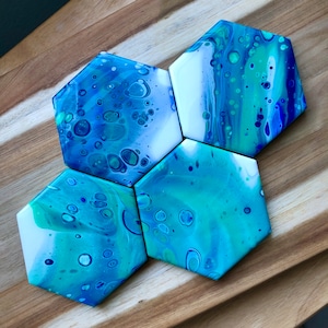 May include: Hexagon-shaped coasters with a fluid art design in shades of blue, teal, and white. The coasters have a glossy finish and are arranged on a wooden surface. The abstract pattern resembles ocean waves.