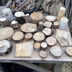May include: A collection of various unfinished wooden discs, blocks, and logs of different sizes and textures. The pieces feature natural bark edges, light brown wood grain, and smooth cut surfaces arranged on a flat wooden surface.