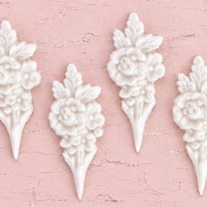 May include: Four identical white floral appliques, each featuring a sculpted bouquet of roses and leaves with a pointed base. These decorative elements have a smooth, matte finish and are arranged in a vertical row against a textured background.
