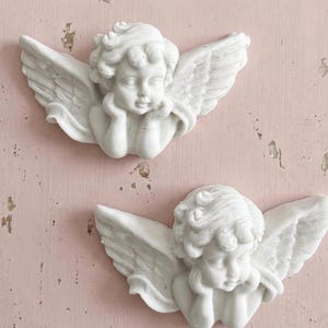 May include: Two white cherub wall plaques featuring sculpted figures of winged infants with curly hair. Each cherub is depicted with hands resting near the chin, showcasing detailed feather textures on the wings and soft facial features in a matte finish.
