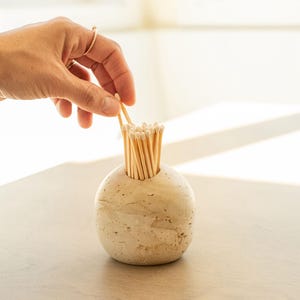 May include: A beige, spherical match holder filled with wooden matches. A hand is reaching in to grab a match. The match holder is made of a stone-like material and sits on a light-coloured surface.