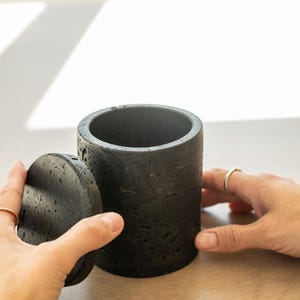 Peut inclure: Un récipient cylindrique en béton gris foncé avec un couvercle assorti. Le récipient a une surface rugueuse et texturée. Le couvercle est légèrement décalé. Design minimaliste et moderne, idéal pour la décoration intérieure.