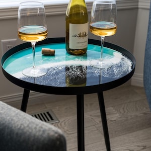 Peut inclure: Une table d'appoint noire avec un plateau rond en résine turquoise et blanche représentant un motif de vague. Deux verres à vin et une bouteille de Chardonnay sont sur la table.
