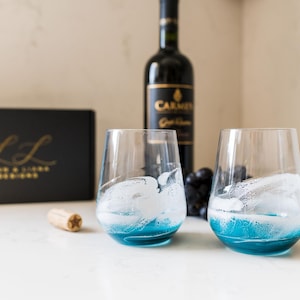 May include: Two blue glass wine glasses with white swirls inside, sitting on a white countertop. A bottle of red wine and a box with the text "L & L Wine & Liquor Boutique" are in the background.