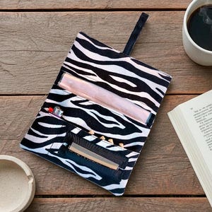 Puede incluir: Un estuche de accesorios con estampado de cebra en blanco y negro con un asa negra, abierto para revelar un encendedor y otros artículos. Una taza de café con vapor, un libro abierto y un cenicero pequeño están sobre una superficie de madera.