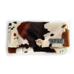 Peut inclure: Un portefeuille rectangulaire avec un motif de peau de vache en fausse fourrure dans des tons marron, noir et crème. Le portefeuille est fermé par un bouton en bois clair et une boucle élastique noire. Une étiquette grise indique BIOTA.BAGS.