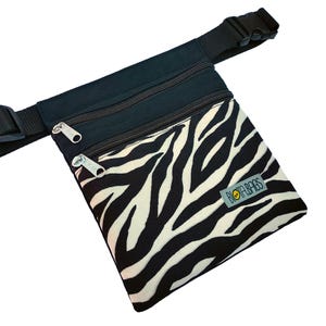 May include: A black waist bag featuring a white and black zebra print on the front pocket. The bag includes two zippered compartments with silver-toned pulls, an adjustable black strap with a plastic buckle, and a label reading BIOTA.BAGS.