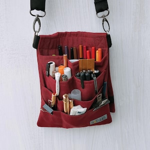 May include: A deep red canvas crossbody organizer bag featuring multiple exterior pockets filled with various pens, markers, and stationery tools. The bag includes a black adjustable shoulder strap with metal clips and a small label reading BIOTA & BAGS.