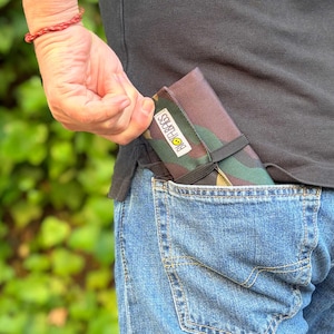 Op de afbeelding: Een camouflage-patroon etui met de tekst "BIOBABES" is gedeeltelijk zichtbaar in een blauwe jeanszak. De etui is vastgezet met een zwarte elastische band. De persoon draagt een zwart shirt.