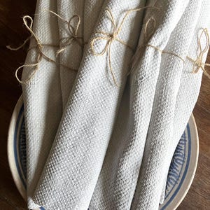 Pode incluir: Um conjunto de guardanapos enrolados em tecido waffle cinza claro, amarrados com laços de barbante, apresentados em um prato de cerâmica com padrões azuis e brancos. Adequado para a mesa.