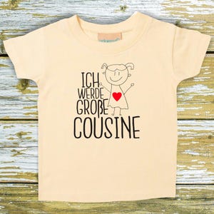 Può includere: Una maglietta giallo chiaro con una grafica nera e rossa che dice "Ich werde große Cousine", che si traduce in "Sarò una grande cugina".