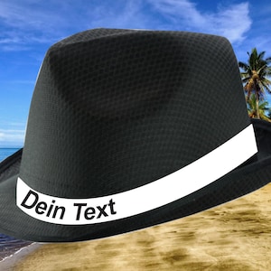 May include: Black fedora hat with a white band and the text "Dein Text" on the band. The hat is on a sandy beach with a blue sea in the background.