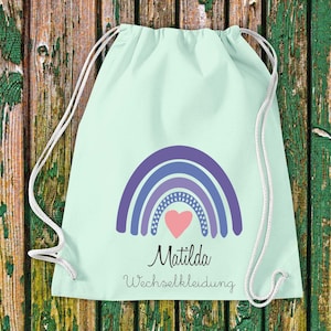 Könnte beinhalten: Eine hellblaue Kordeltasche mit einem Regenbogenmuster und einem rosa Herz in der Mitte. Die Tasche trägt den Text "Matilda Wechselkleidung".