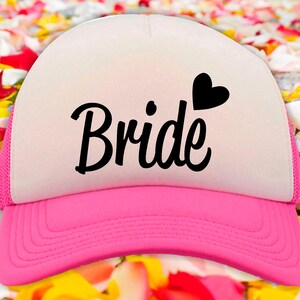 Peut inclure: Un chapeau de camionneur blanc et rose avec un filet noir à l'arrière. Le devant du chapeau porte le mot "Bride" écrit en cursive noire avec un petit cœur noir au-dessus.