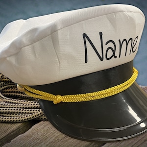 Könnte beinhalten: Ein weißer Kapitänshut mit schwarzem Rand und gelber Kordelverzierung. Der Hut trägt den Schriftzug "Name" in Schwarz auf der Vorderseite.