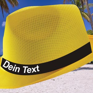 May include: A yellow straw hat with a black band and the text "Dein Text" on the band. The hat is sitting on a sandy beach with palm trees in the background.