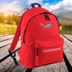 May include: Red backpack with a cartoon wolf graphic. The backpack has a front pocket with the word "Wunschname" in white script. Features black zippers and straps. The background shows a lake and mountains.