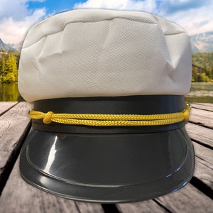 May include: A white captain's hat with a black brim and a yellow rope trim. The hat is on a wooden surface.
