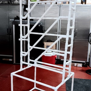 May include: White PVC pipe frame with a rectangular base. The frame is made of multiple pieces of pipe connected together to form a tall, open structure.