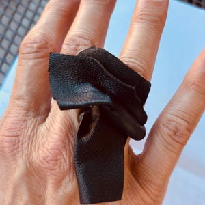 Op de afbeelding: Een zwarte leren ring met een getextureerd oppervlak. De ring is gemaakt van gevouwen leer en heeft een uniek, artistiek ontwerp. De ring is een opvallend sieraad.