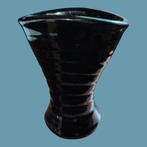 May include: A glossy black ceramic vase with a flared, triangular-shaped rim and a narrow, cinched neck that widens toward the base. The exterior surface features a series of horizontal, undulating ridges that create a textured, rippled appearance.