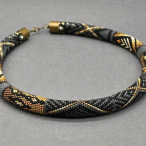 May include: A beaded necklace in a tubular design, featuring a pattern of black, gray, and gold beads. The necklace has a clasp closure and a mix of geometric and linear patterns.