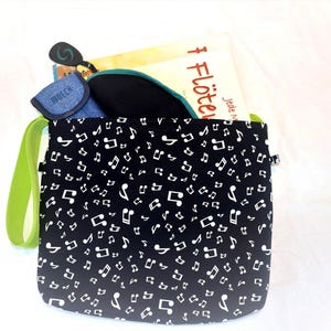 Könnte beinhalten: Eine schwarze Tragetasche mit weißem Notenmuster. Die Tasche hat einen grünen Riemen und enthält ein Buch, ein blaues Etui mit dem Wort "MOECH" und andere Gegenstände. Das Buch trägt den Titel "Flöten".