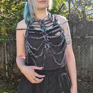 May include: A black and iridescent crochet top with a halter neck design. The top features a series of connected chains that create a cage-like effect. The crochet work incorporates shades of black, blue, and purple.