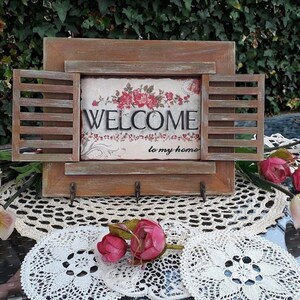 May include: A wooden sign with a window-like design and a welcome message. The sign has a rustic brown finish and features a white background with red roses and the text "WELCOME to my home".