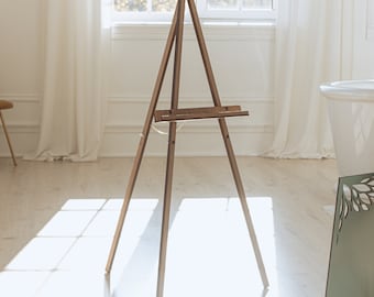 Puede incluir: Un caballete de madera de tres patas con un acabado marrón medio. La estructura cuenta con un soporte vertical central, dos patas delanteras inclinadas y un estante horizontal para colocar objetos, unido por una fina cadena de metal.