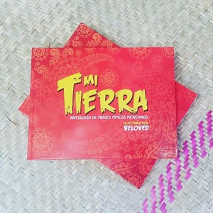 May include: A red book titled "Mi Tierra" with yellow text, featuring an anthology of Mexican traditional costumes. The cover has a floral pattern and the text "Ilustrado por Beloved". The book is on a woven surface.