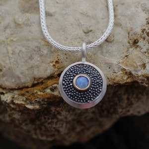 Könnte beinhalten: Eine silberne Kettenkette mit einem runden Anhänger, der einen blauen Opal-Stein in einer silbernen Fassung mit goldener Einfassung trägt. Der Anhänger ist von einer strukturierten silbernen Oberfläche umgeben.