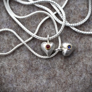Può includere: Due ciondoli a forma di cuore in argento con pietre preziose rosse e blu appesi a una catena d'argento. La catena è appoggiata su una superficie grigia e pelosa.