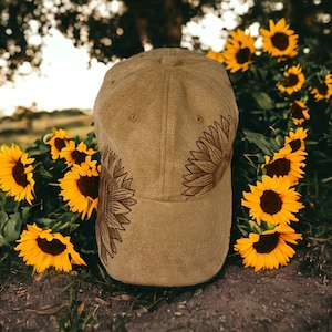 Può includere: Un cappello da baseball marrone con girasoli ricamati sul davanti e sul lato. Il cappello è appoggiato a terra davanti a un campo di girasoli.