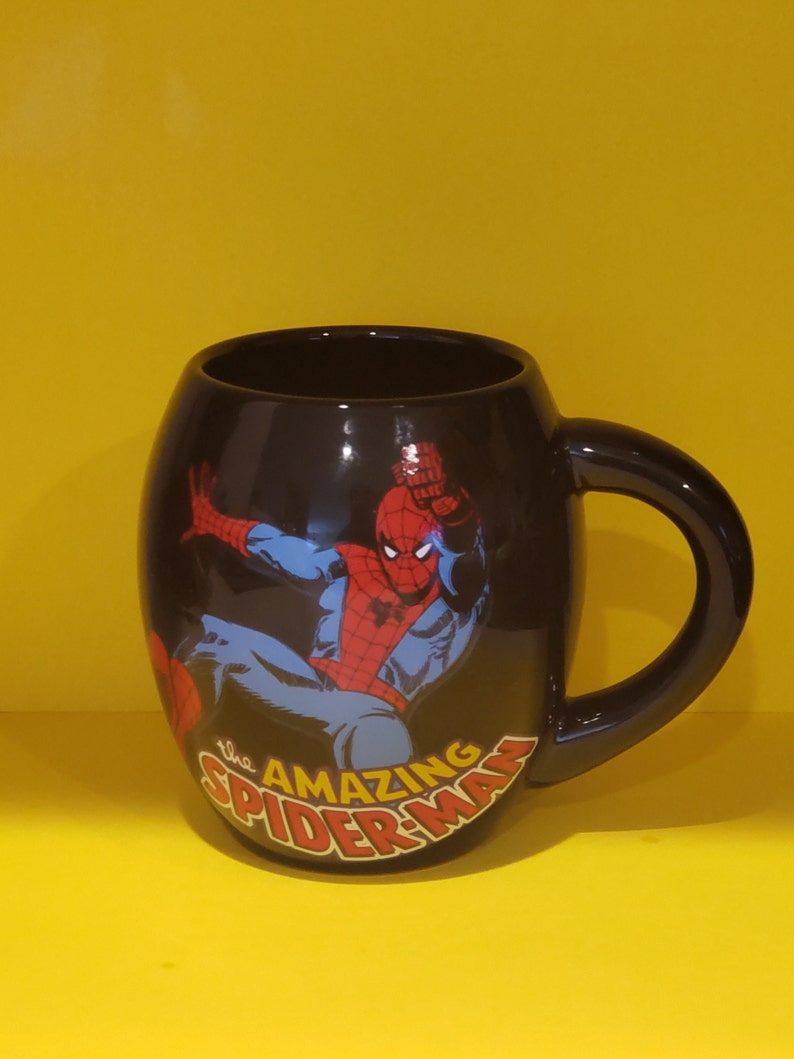 May include: A black ceramic mug with a graphic of Spider-Man in a red and blue suit. The mug has the text "The Amazing Spider-Man" printed on the front.