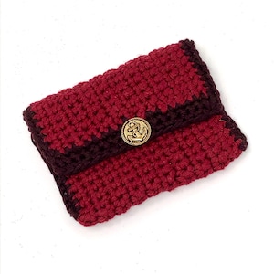 Può includere: Un piccolo portafoglio all'uncinetto di colore bordeaux e marrone scuro. Il portafoglio ha una chiusura a bottone con un'ancora dorata.