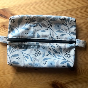 Könnte beinhalten: Eine kleine, mit einem Reißverschluss versehene Tasche mit einem Blumenmuster in Blau- und Grautönen. Die Tasche hat einen schwarzen Reißverschluss und ein kleines Etikett mit der Aufschrift "KOJARI".