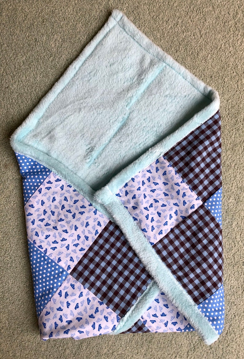 Puede incluir: Una manta de beb&eacute; de patchwork azul y marr&oacute;n con un forro de felpa azul claro y suave. La manta est&aacute; hecha de diferentes telas con varios patrones, incluyendo lunares, cuadros vichy y un peque&ntilde;o estampado de barcos.