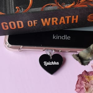 Puede incluir: Un dije negro en forma de corazón con el nombre "Lisichka" en letras blancas cuelga de un dispositivo Kindle. El libro "God of Wrath" es visible, junto con rosas rosas secas sobre un fondo rosa.