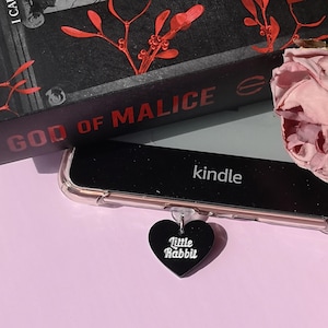 May include: A black Kindle e-reader in a clear case, with a black heart-shaped charm that reads "Little Rabbit." A book titled "God of Malice" is visible in the background, along with dried roses. The Kindle is on a pink surface.