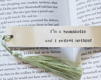May include: A wooden bookmark with a light brown finish and a green tassel. The bookmark has the text "i'm a bookaholic and i regret nothing" printed on it.