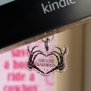 May include: A clear acrylic charm with a skeleton hand design holding a heart, with the words "TRIGGER WARNINGS" in black. The charm is attached to a Kindle e-reader. The background is a blurred pink color.
