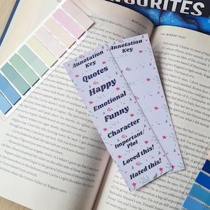 May include: Two purple bookmarks with floral designs and white text. The bookmarks are labelled "Annotation Key" and feature categories for quotes, happy, emotional, funny, character/important plot, loved this, and hated this.