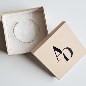 May include: A silver bangle bracelet in a brown cardboard box with the letters "AD" printed on the top of the box.
