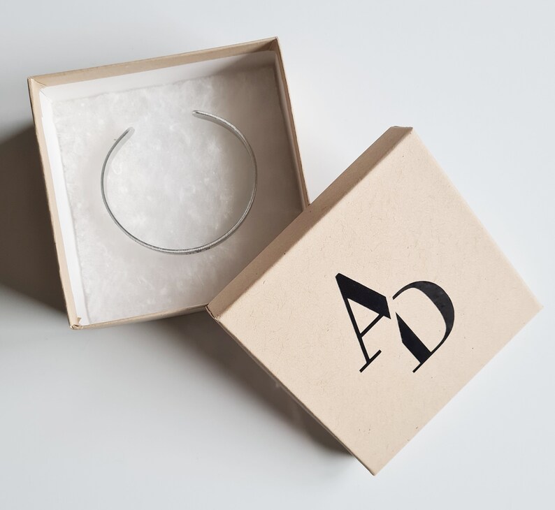 May include: A silver bangle bracelet in a brown cardboard box with the letters "AD" printed on the top of the box.