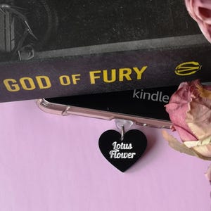 May include: A black heart-shaped charm with the words "Lotus Flower" in white script. The charm is attached to a clear phone case, with a book titled "God of Fury" in the background. A pink flower is in the corner.