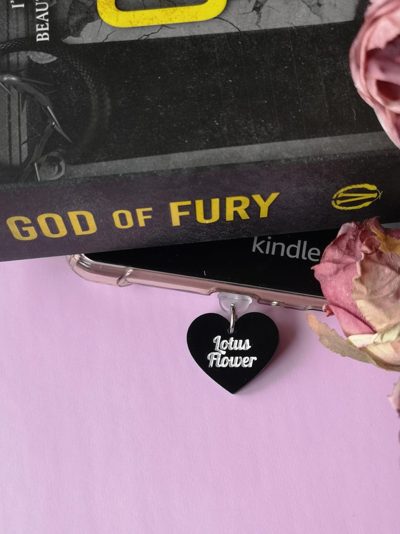 May include: A black heart-shaped charm with the words "Lotus Flower" in white script. The charm is attached to a clear phone case, with a book titled "God of Fury" in the background. A pink flower is in the corner.
