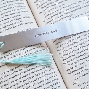 May include: A silver metal bookmark with a light blue tassel. The bookmark has the text "your text here" engraved on it.
